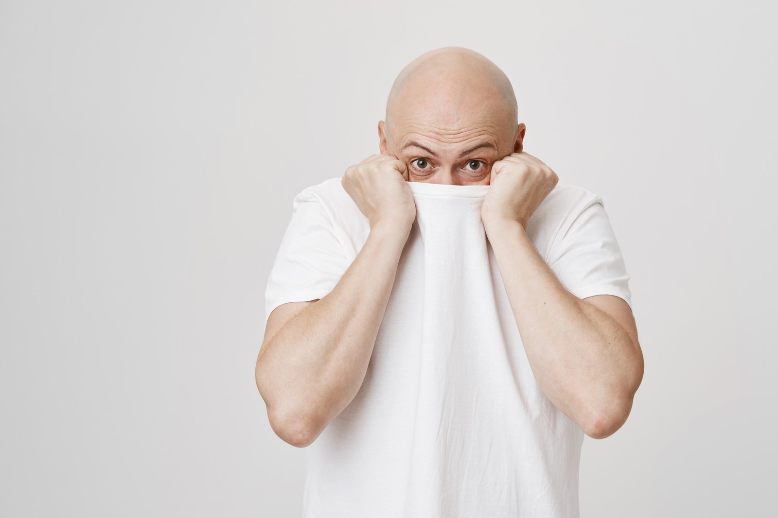 Indoor shot of funny bald caucasian man hiding head in his white t-shirt and glancing at camera
